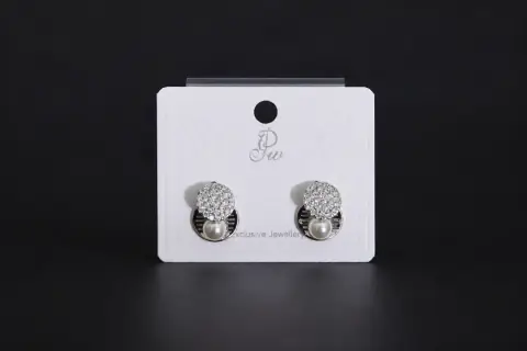 Pair of silver-toned geometric stud earrings featuring a round crystal pave disc and a small white pearl drop on a display card.